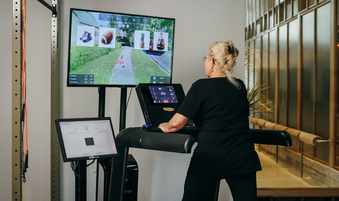 An older adult using a treadmill with GaitBetter's motor-cognitive training system, interacting with a virtual environment displayed on a screen.