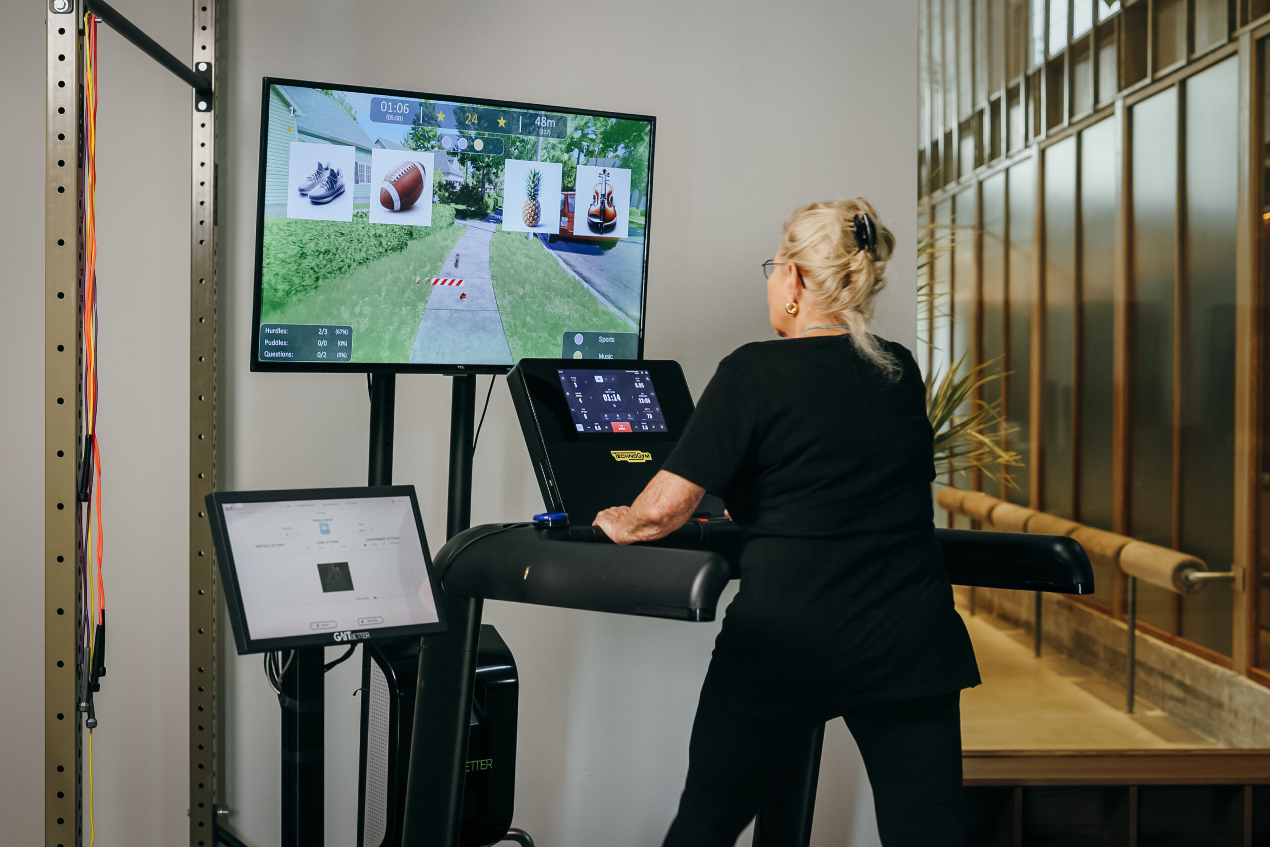 An older adult using a treadmill with GaitBetter's motor-cognitive training system, interacting with a virtual environment displayed on a screen.