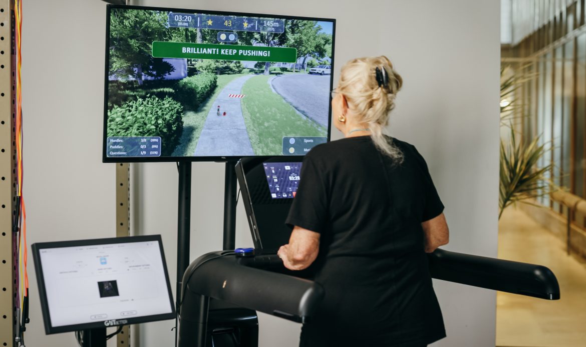 GaitBetter system in action, on a TV screen, with a lady walking on a treadmill, while looking at the TV screen.