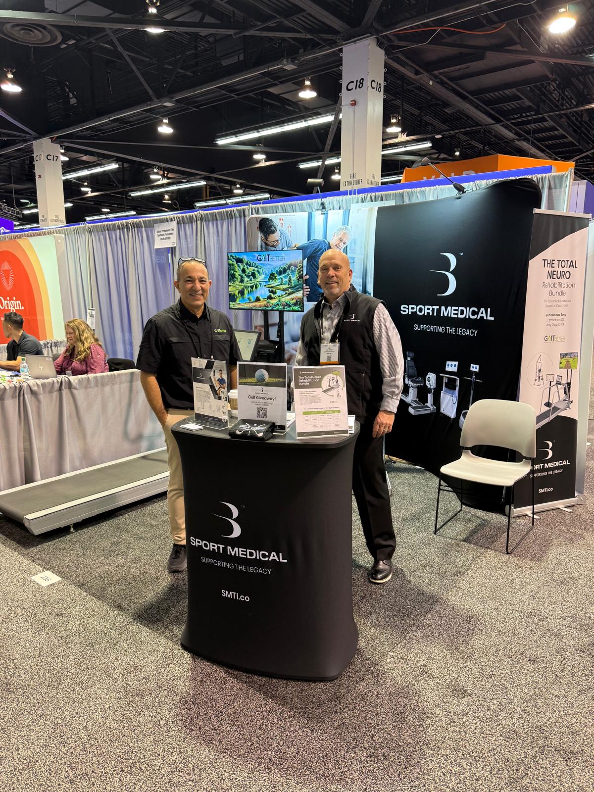 Two men standing at an exhibition booth, they are smiling at the camera. The booth has branded signage for Sport Medical, and GaitBetter. There is a treadmill with GaitBetter on the TV screen next to the treadmill.